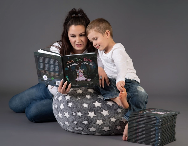 A parent sits on a plush rug, reading a children's book titled "One, Two, Three, Meditate with Me" to a young child.